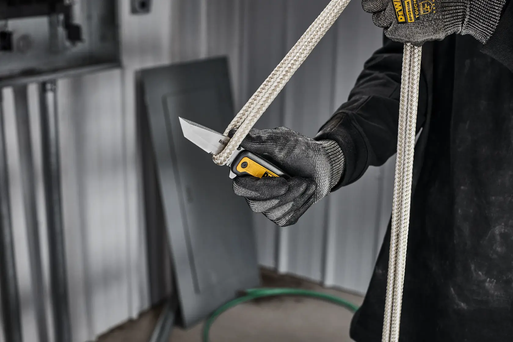 A person wearing gloves is using a DEWALT folding knife to cut a thick rope in an industrial setting.