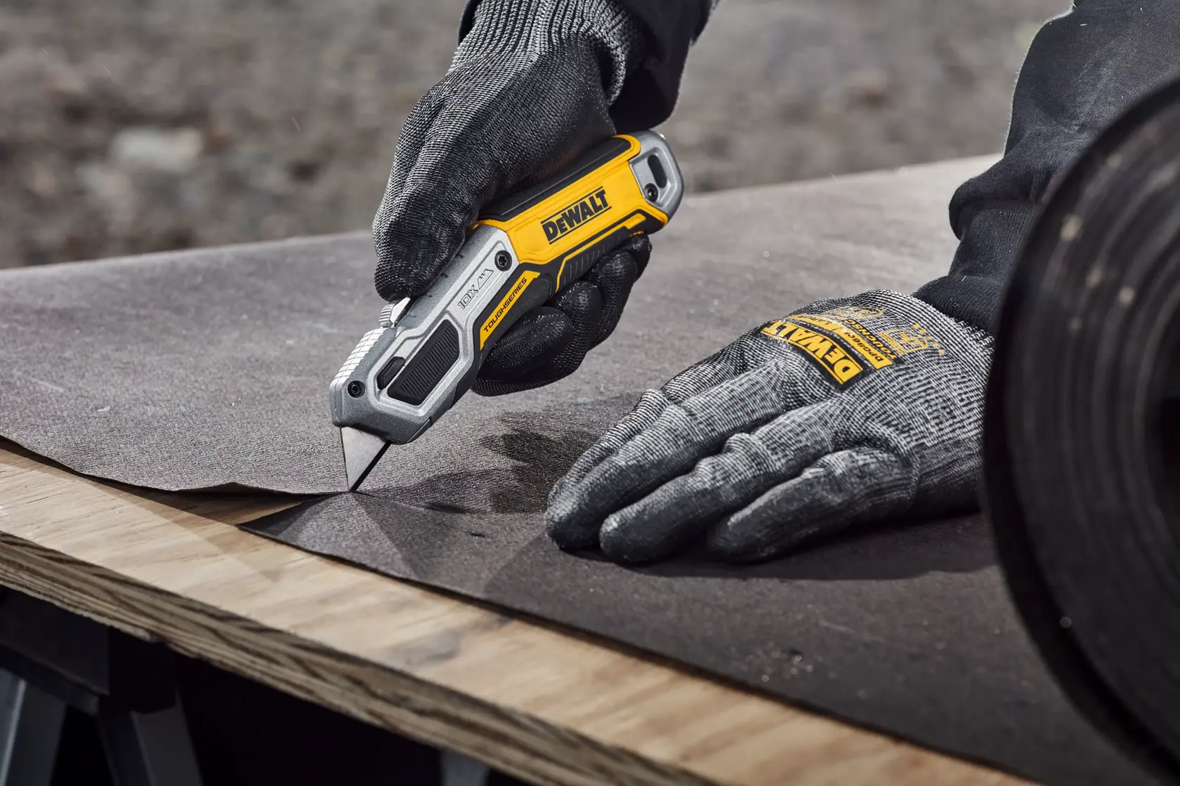 A person wearing protective gloves is using a DEWALT utility knife to cut a sheet of material on a wooden workbench.