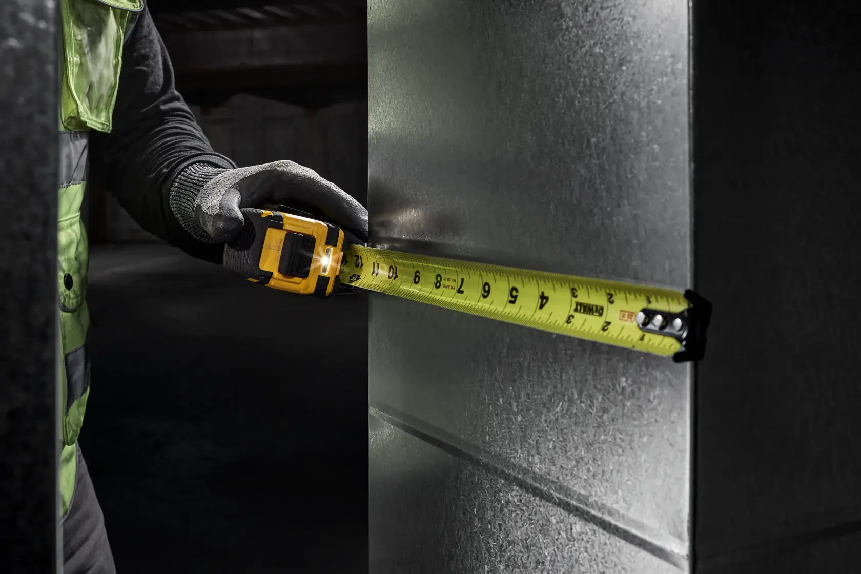 A person wearing gloves is using a yellow DEWALT tape measure to measure the width of a metallic surface.