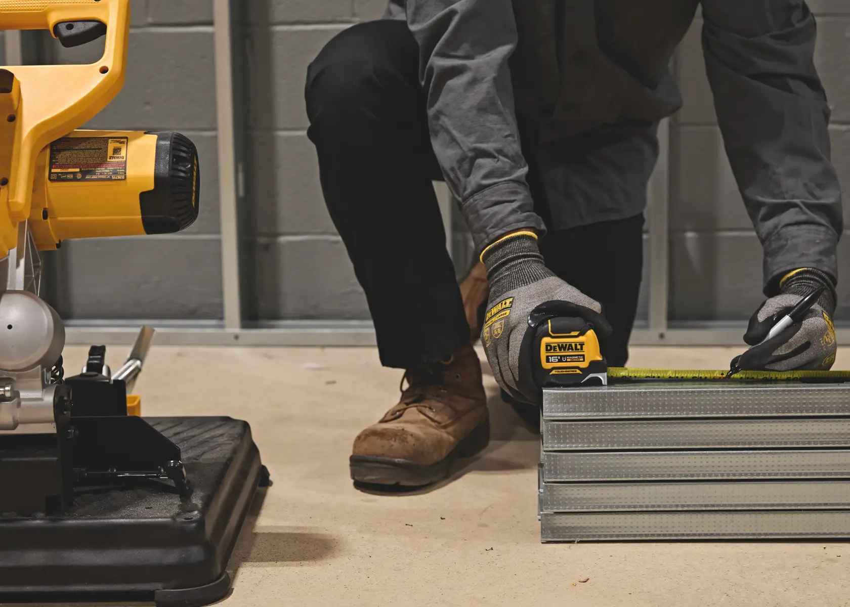 A person wearing work gloves and boots uses a DEWALT tape measure to mark measurements on metal building studs. The scene also includes part of a yellow DEWALT power tool in the background on the left side.