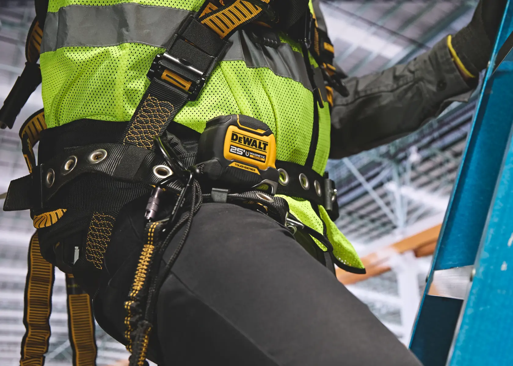 Close-up of a person wearing a safety harness and a high-visibility vest, climbing a blue ladder. A DEWALT 25 ft tape measure is attached to their belt. The background shows an indoor industrial setting.