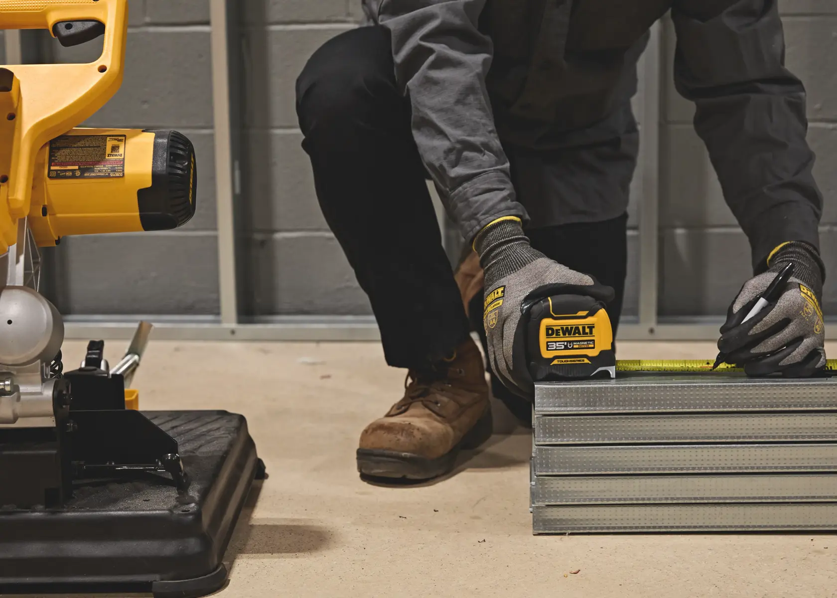 A person wearing work gloves uses a DEWALT tape measure to mark measurements on metal studs in a workshop, with construction equipment visible nearby.