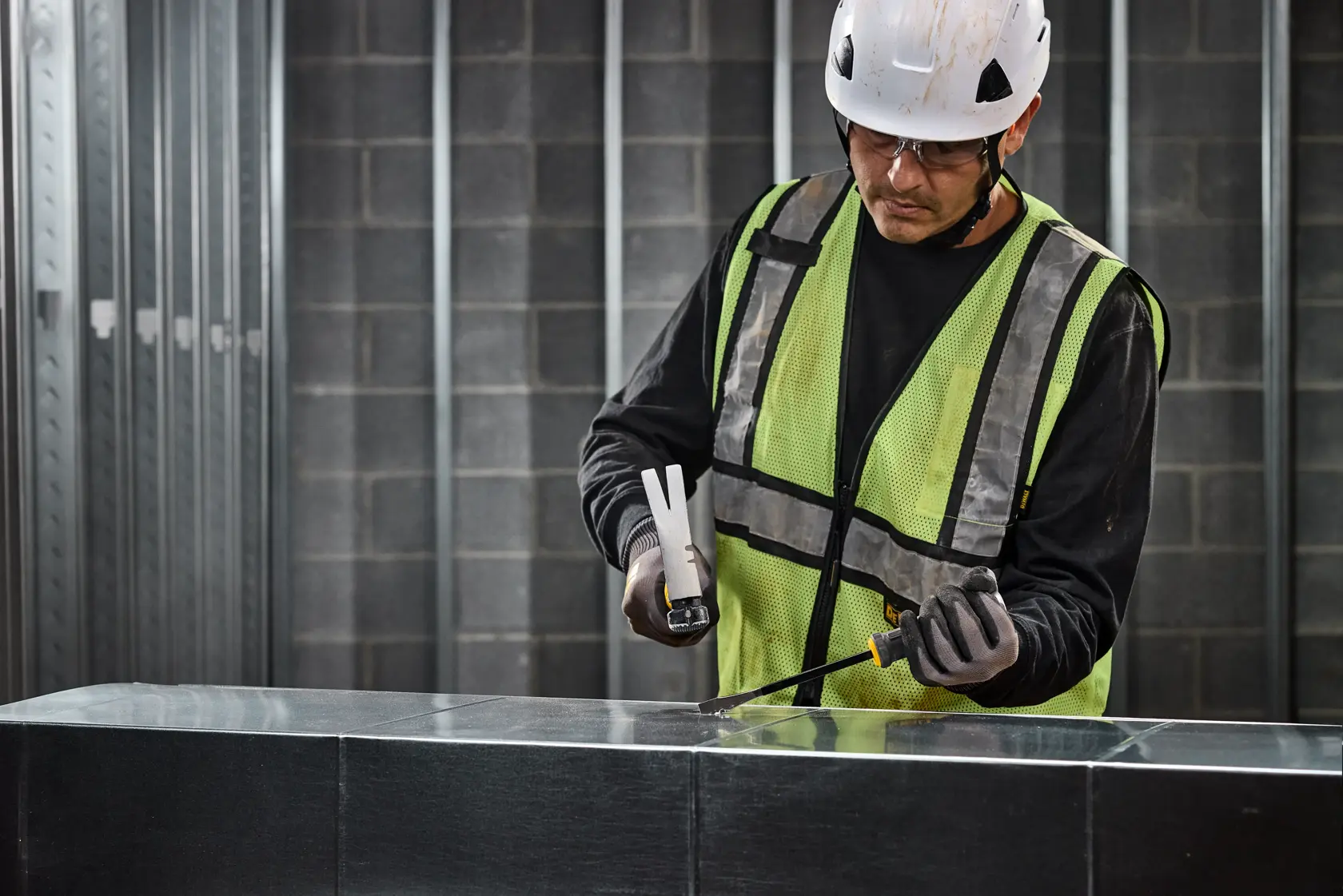 A person wearing a reflective safety vest, gloves, and a hard hat is using a DEWALT bar tool (SKU: DWHT65106) on a metal surface at an industrial worksite. The face of the person is blurred for privacy.