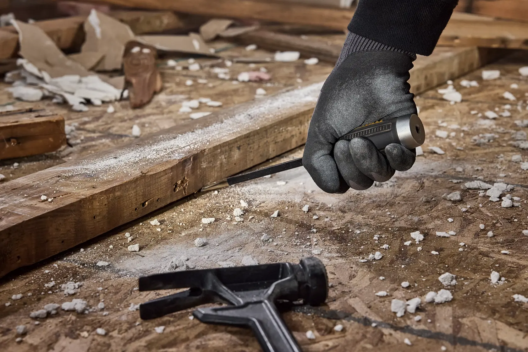 A gloved hand uses a DEWALT screwdriver to work on a wooden plank amidst debris in a construction or renovation setting. A black pry tool is also visible on the dusty wooden surface.