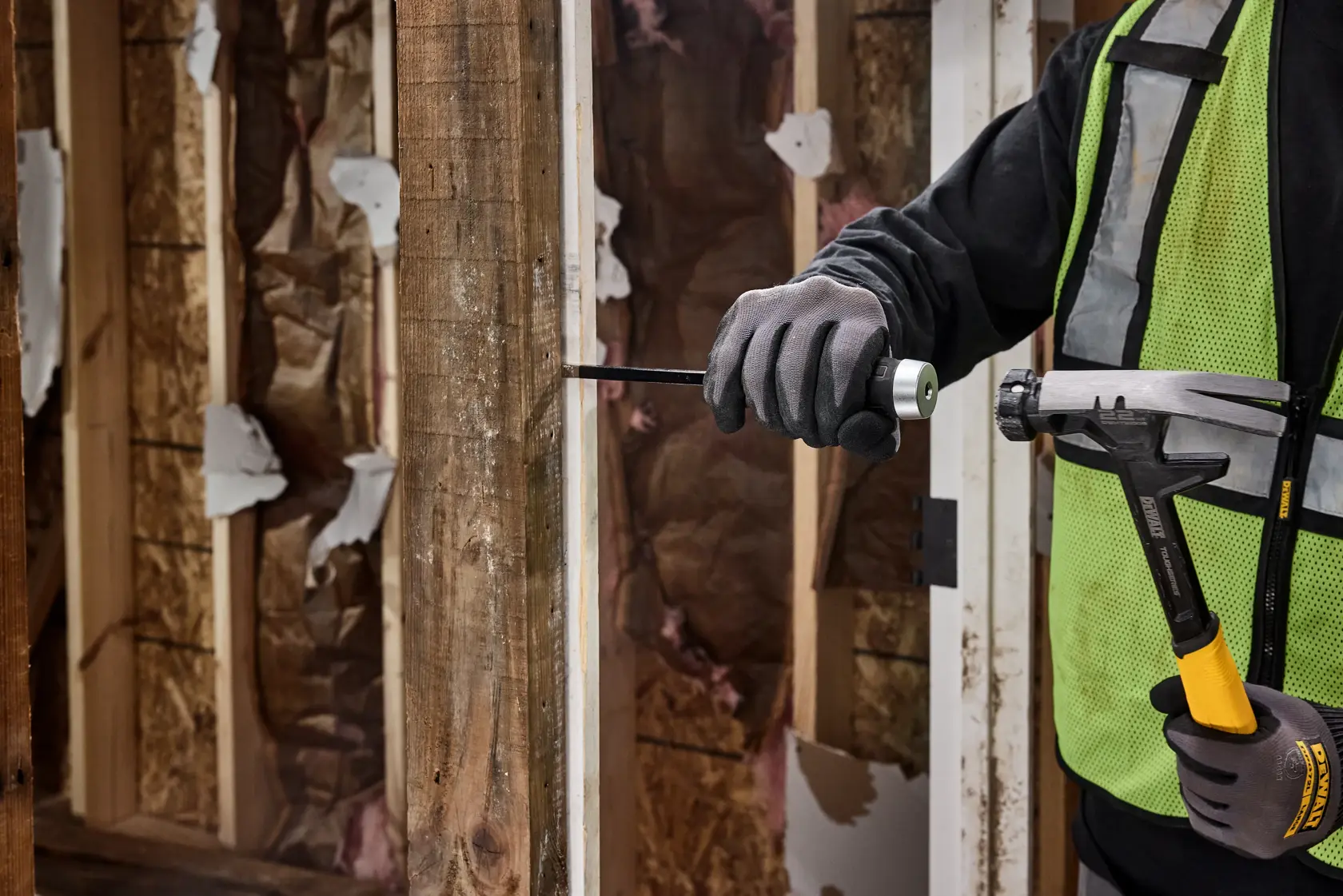 A person wearing construction gloves and a safety vest is using a DEWALT hammer and a screwdriver to work on a wooden frame at a construction site.