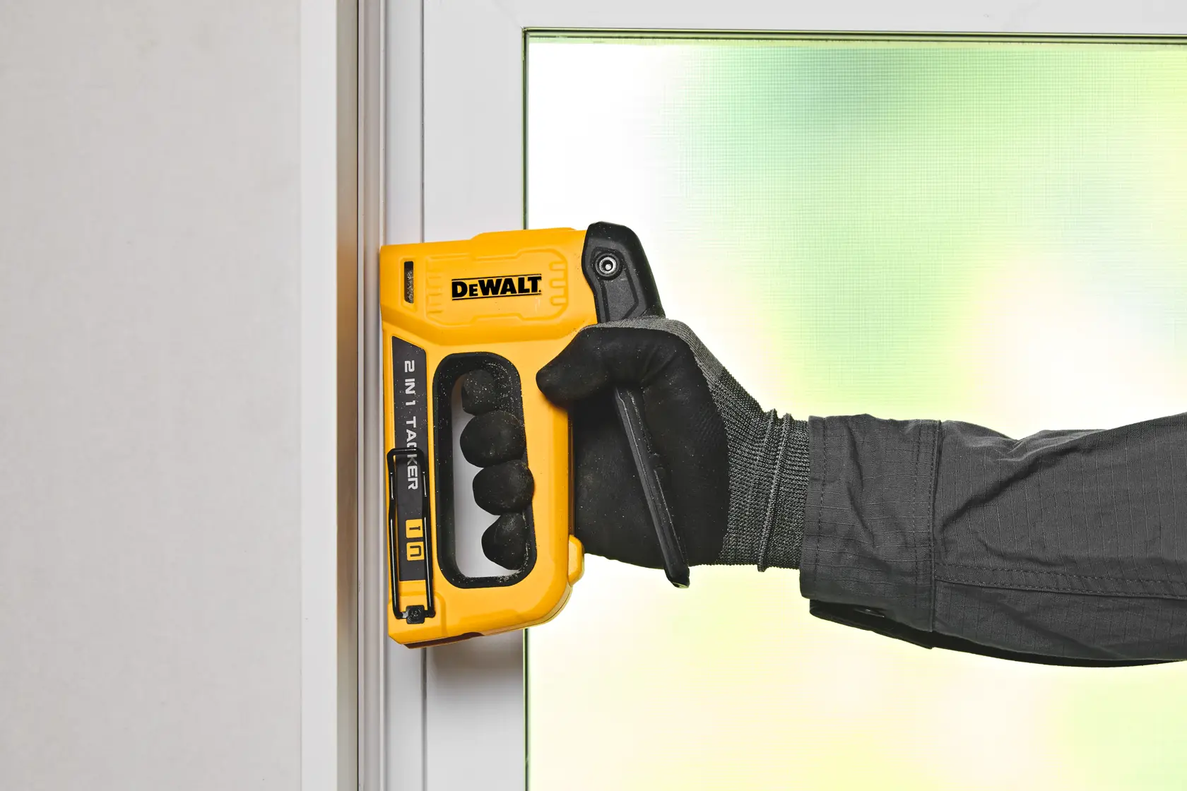 A person wearing black gloves is using a yellow DeWalt 2-in-1 stapler and nail gun tool on a white window or door frame.