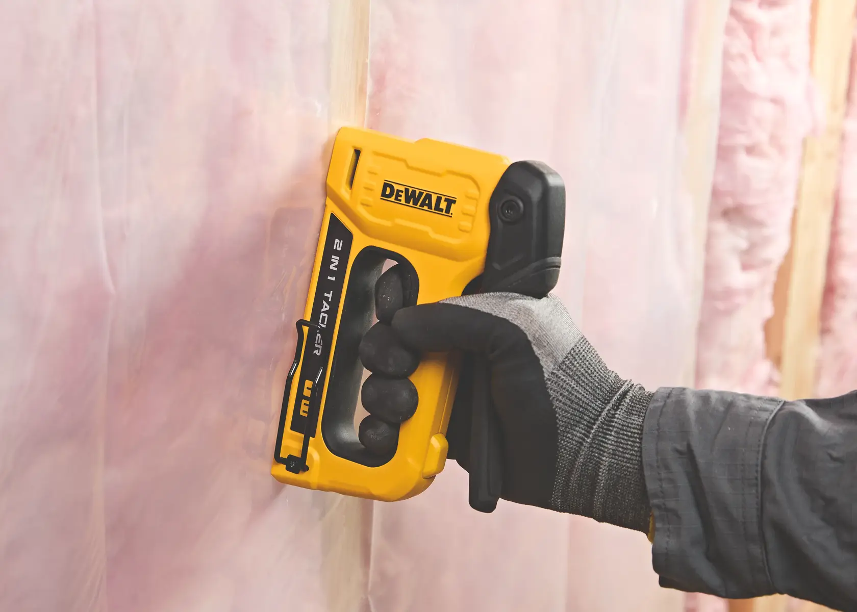 A person wearing a work glove is using a yellow and black DEWALT 2-in-1 tacker stapler to fasten clear plastic sheeting to a surface, with pink insulation visible in the background.