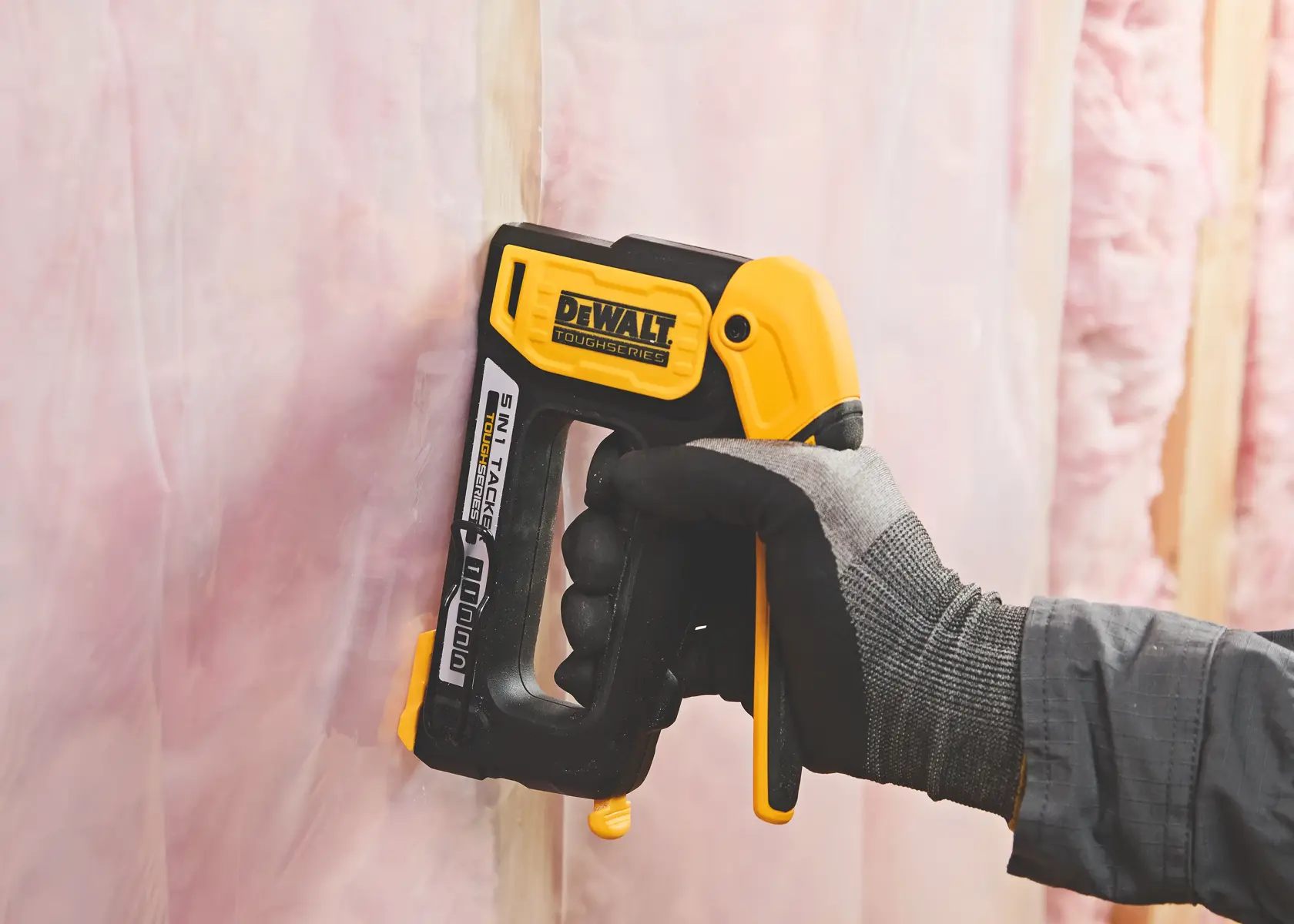 A person wearing a glove is using a DEWALT TOUGHSERIES 5-in-1 manual staple gun to fasten a plastic sheet to insulation material on a wall.