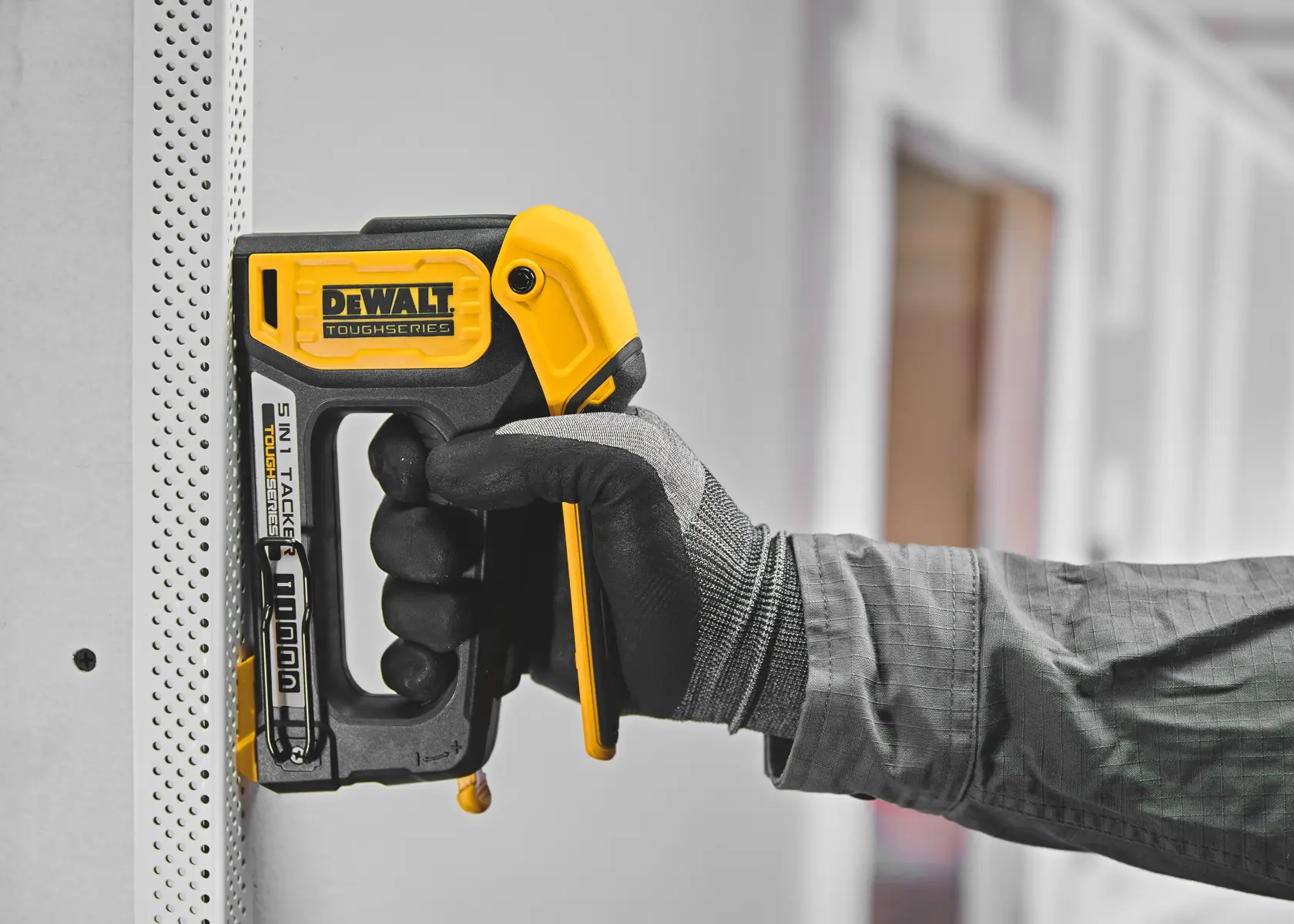 A person wearing a work glove uses a yellow and black DeWalt ToughSeries 5-in-1 tacker tool to staple into a perforated panel indoors.