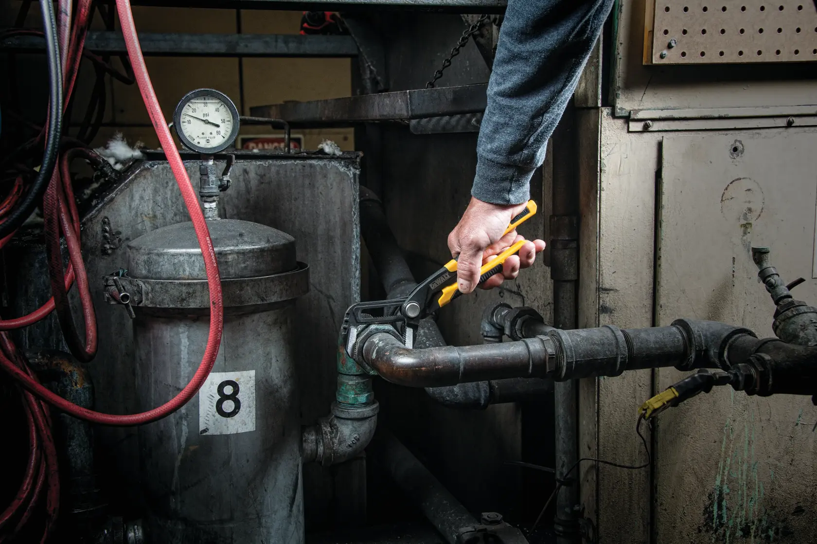 A person uses a DEWALT yellow-handled adjustable wrench (SKU: DWHT74427) to tighten a pipe fitting in an industrial setting, surrounded by metal pipes, a pressure gauge, and machinery.