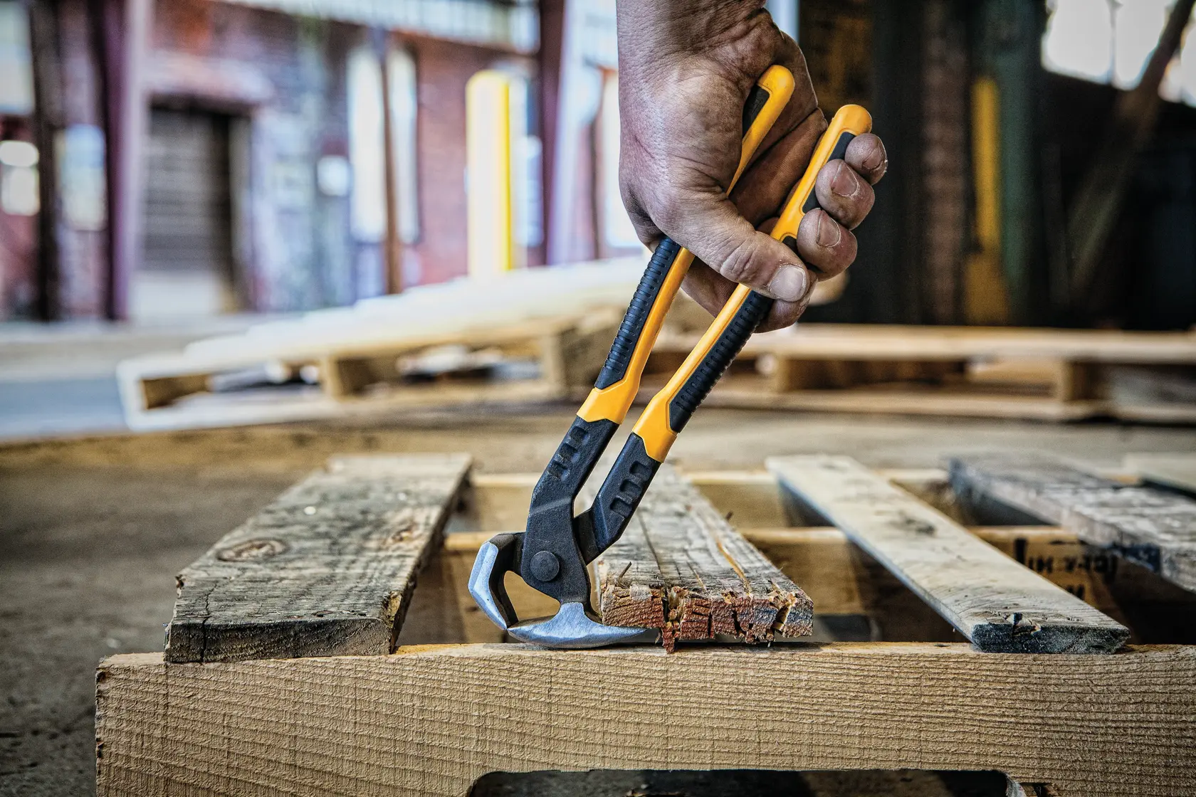 A close-up of a hand using a DEWALT yellow and black hand tool to grip and remove nails from a wooden pallet in an industrial setting.