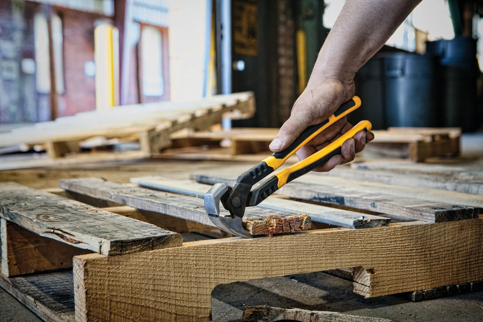 A person uses a DEWALT yellow and black tool, likely a nail puller or pliers, to remove nails from a wooden pallet in an industrial setting.