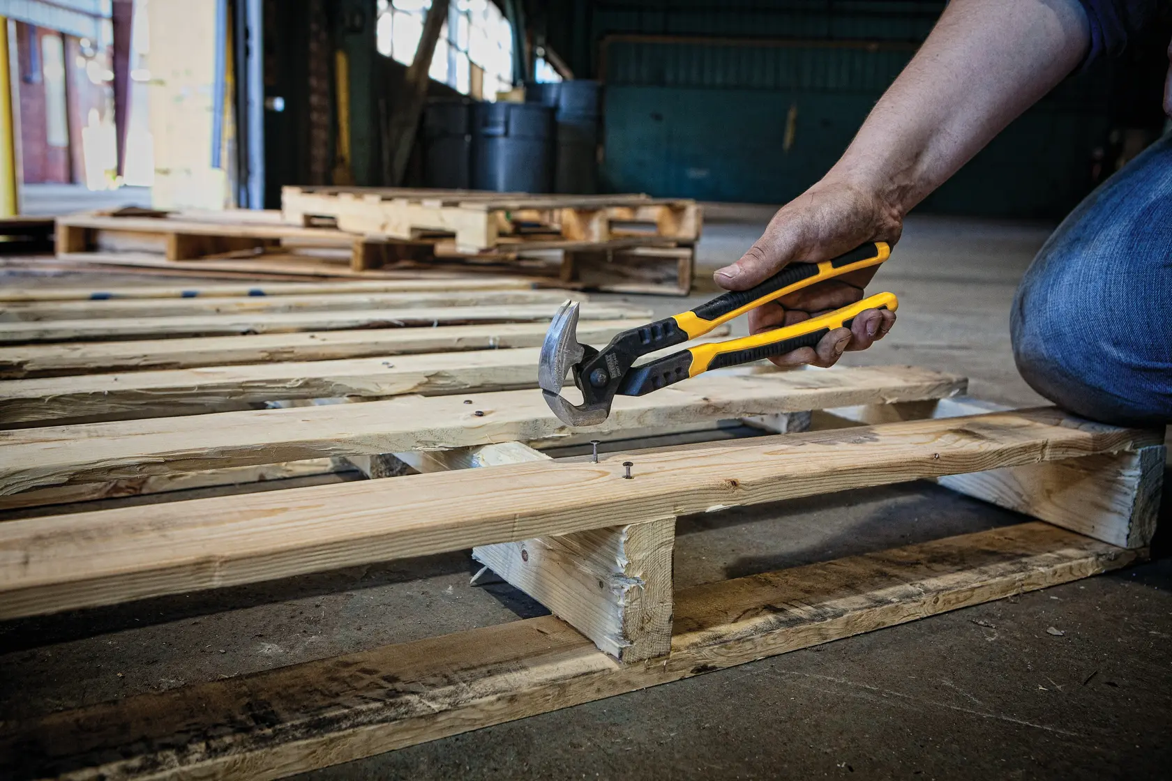 A person uses a DEWALT yellow and black cutting tool to remove nails from a wooden pallet in an industrial setting.