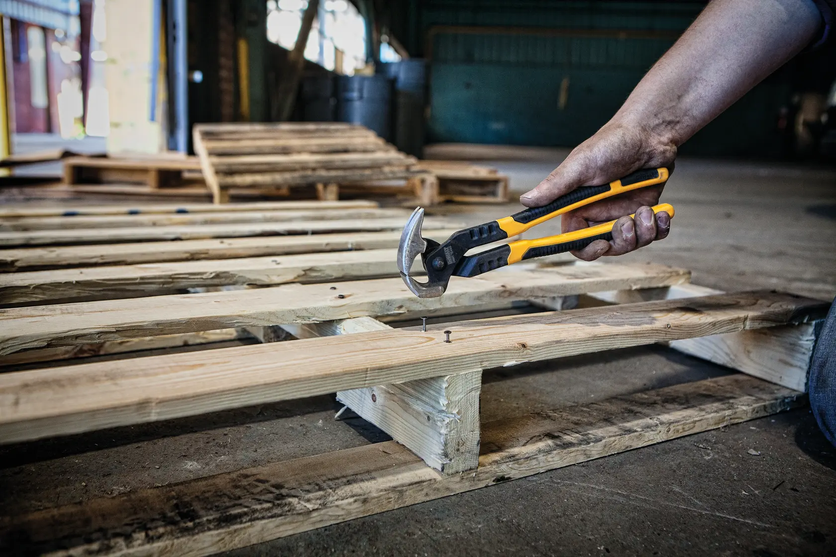 A person using a DEWALT claw bar to remove nails from a wooden pallet inside a workshop.