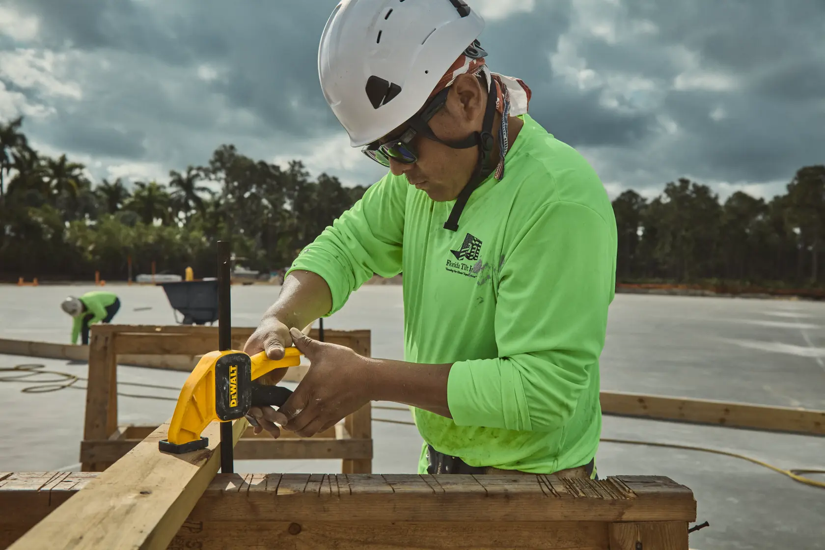 A construction worker wearing a safety helmet and neon green shirt uses a yellow DEWALT clamp to secure wooden beams at an outdoor job site.