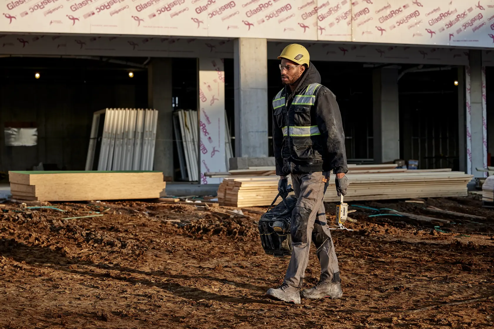 A construction worker wearing a yellow helmet, reflective vest, and work clothes walks across a muddy construction site, carrying a DEWALT tool and a black tool bag. The background shows an unfinished building with stacks of construction materials.