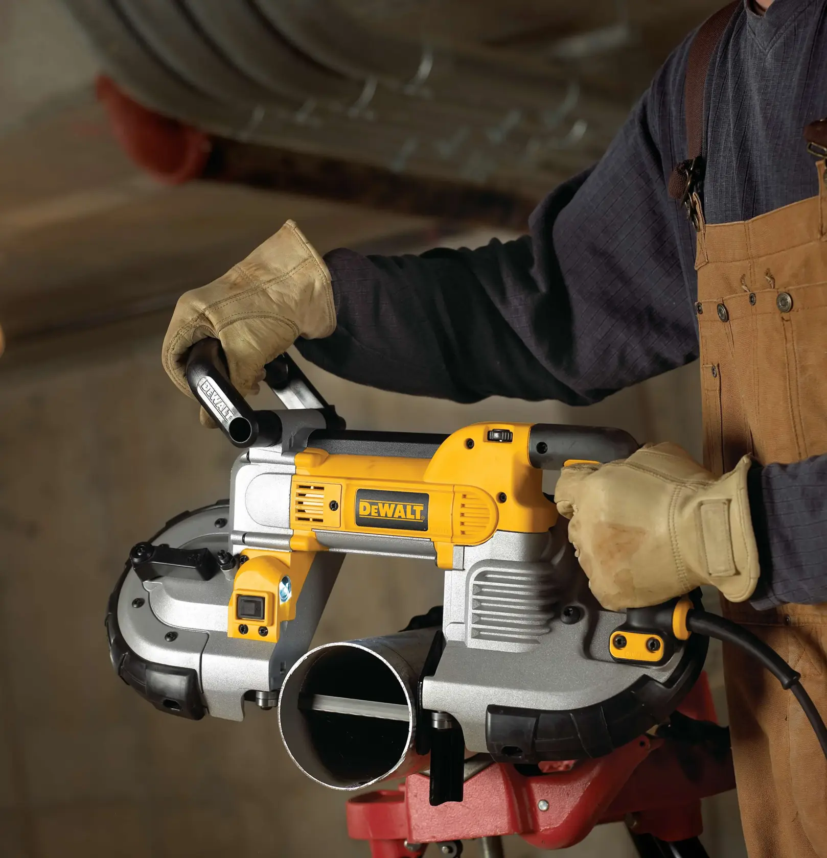 A person wearing gloves and a brown work apron is operating a DEWALT portable band saw, cutting through a metal pipe in an industrial setting.