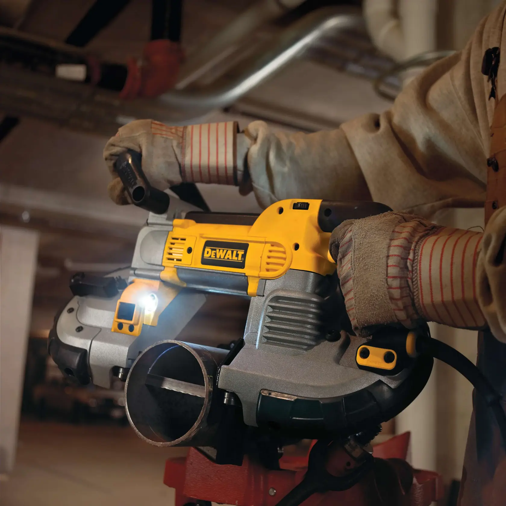 A person wearing protective gloves and clothing is using a DEWALT portable band saw to cut a metal pipe indoors. The tool is powered and a built-in light is illuminating the cutting area.