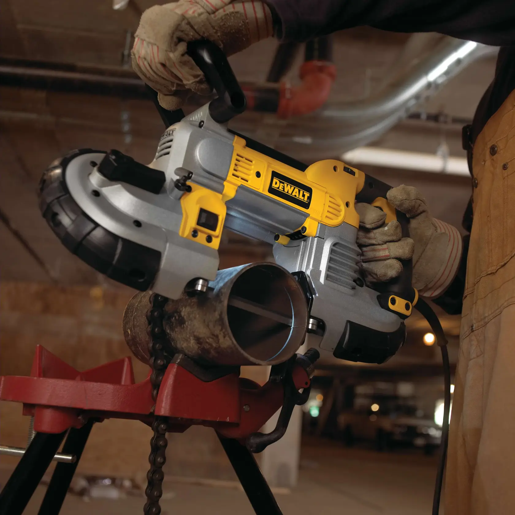 A person wearing work gloves uses a DEWALT portable band saw to cut a metal pipe secured in a red vise stand in an industrial setting.