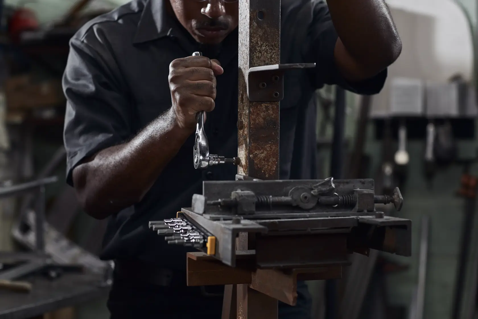 A person wearing a dark shirt is using a DEWALT ratcheting wrench on a metal workpiece held in a vise in a workshop environment. The person's face is blurred, and various tools and equipment are visible in the background.