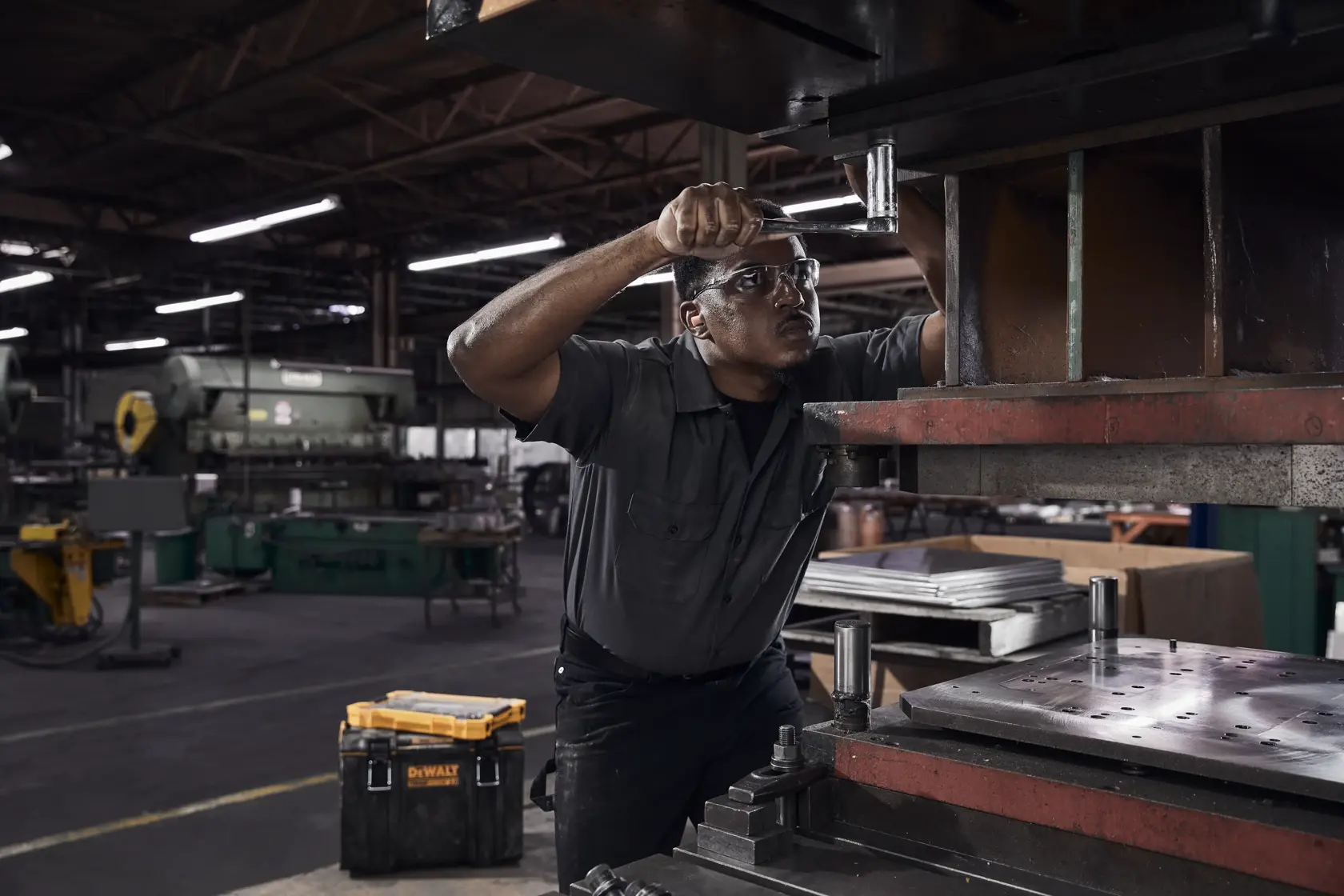 A worker wearing a dark uniform is operating a large industrial machine in a factory setting. There is a yellow tool box labeled 'DeWalt' on the floor beside him, and various machinery and equipment are visible in the background.