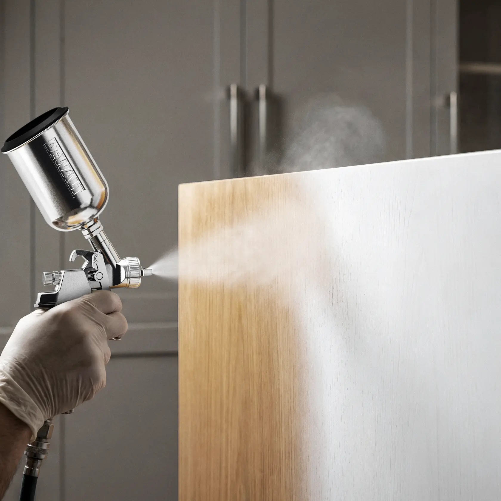 A hand wearing a glove uses a DEWALT spray gun to apply a white coating onto a wooden surface, partially covering the wood grain. The background shows a blurred cabinet.
