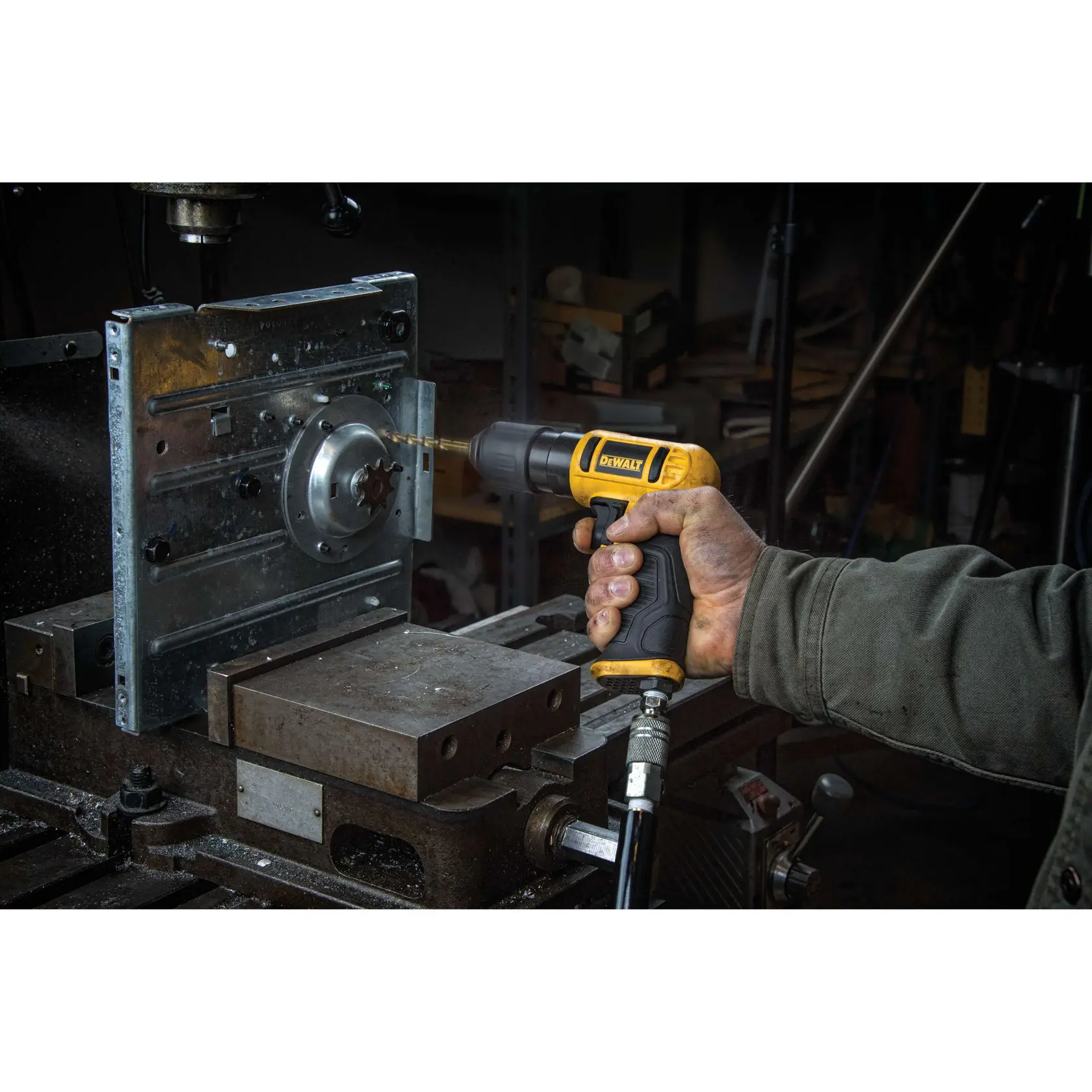 A person is using a DEWALT pneumatic drill to bore into a metal workpiece mounted in a workshop setting. The drill is connected to an air hose and the workspace features industrial equipment.