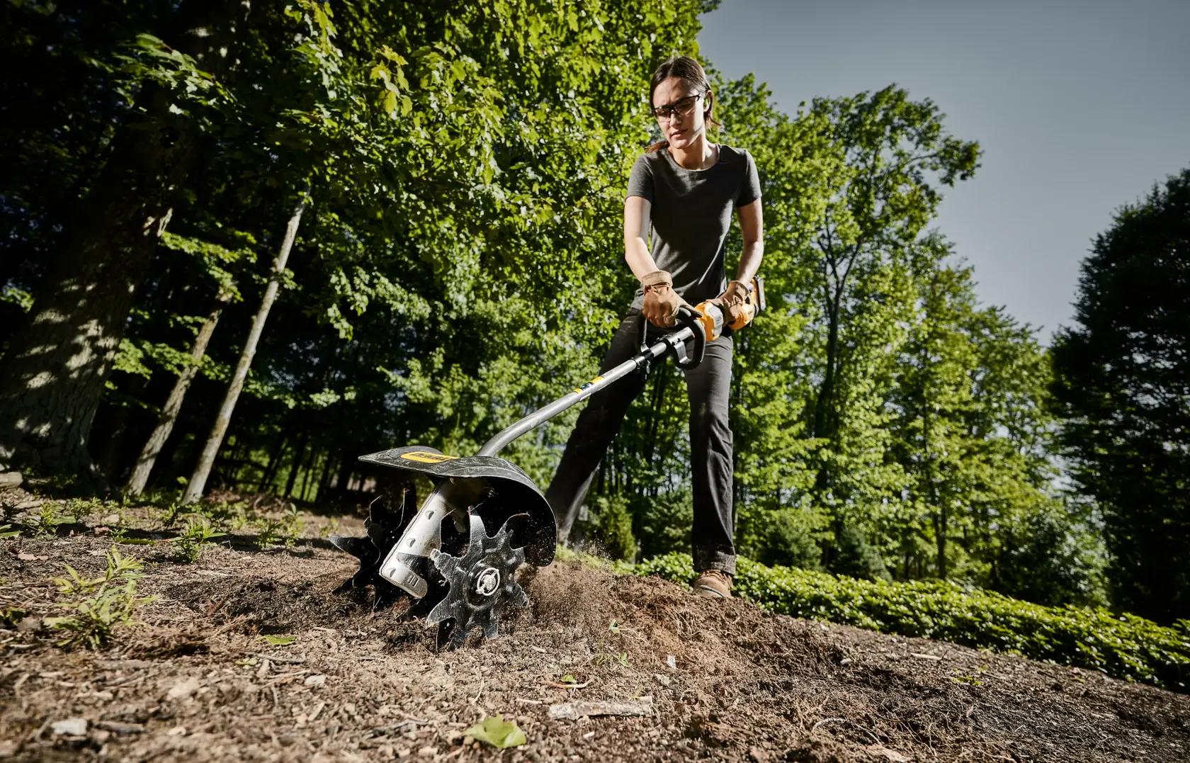 DEWALT cultivator attachment head being used be a woman to work soil in a yard viewed from right angle below. 
