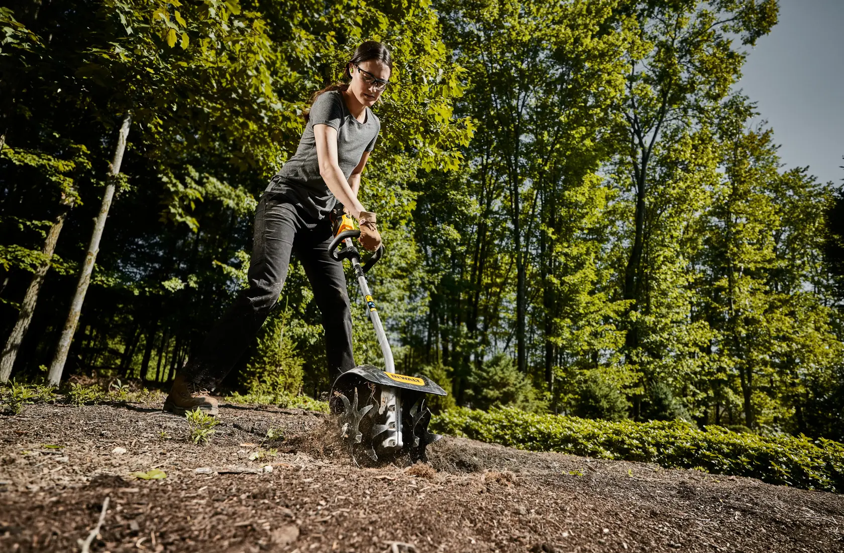 DEWALT cultivator attachment head being used be a woman to work soil in a yard viewed from left angle below. 