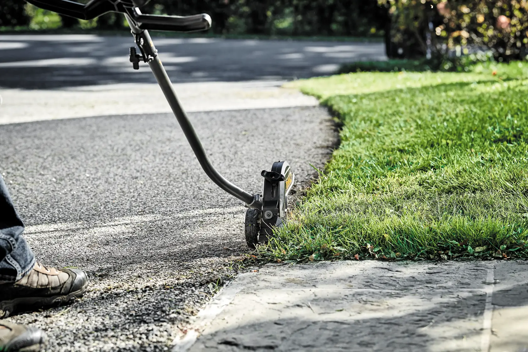A DEWALT lawn edger tool trimming the edge of the grass along a driveway, operated by a person wearing outdoor shoes.