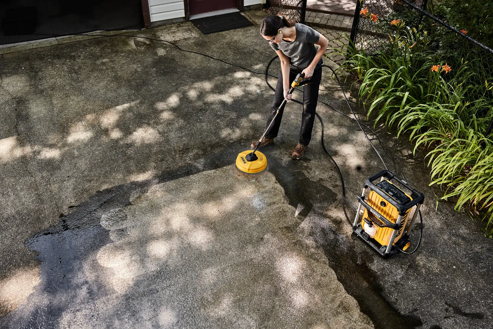 DEWALT 15 Amp Electric Cold Water Pressure Washer being used by a woman cleaning concrete with surface cleaner attachment from birdseye view. 
