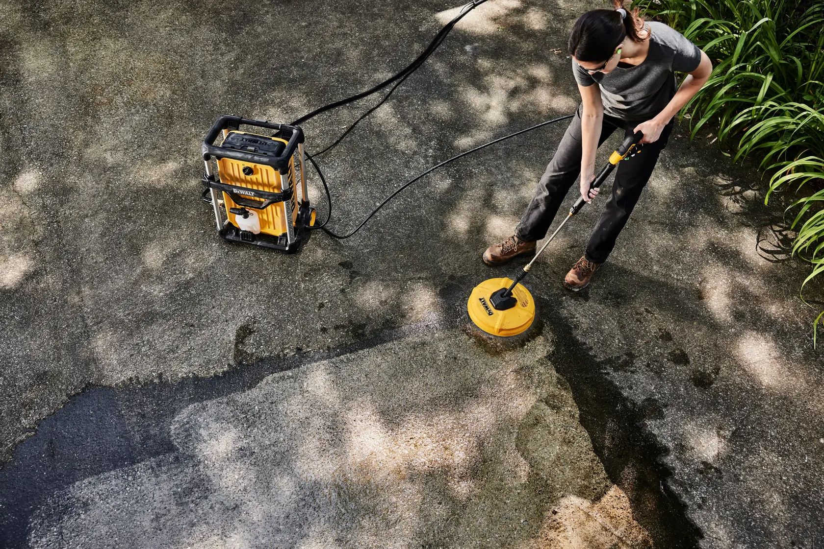 A person is cleaning an outdoor surface with a DEWALT pressure washer (model DWPW3000) and a yellow surface cleaning attachment. The pressure washer and cleaning tool are connected by hoses, and the cleaned section of the ground is visibly lighter.