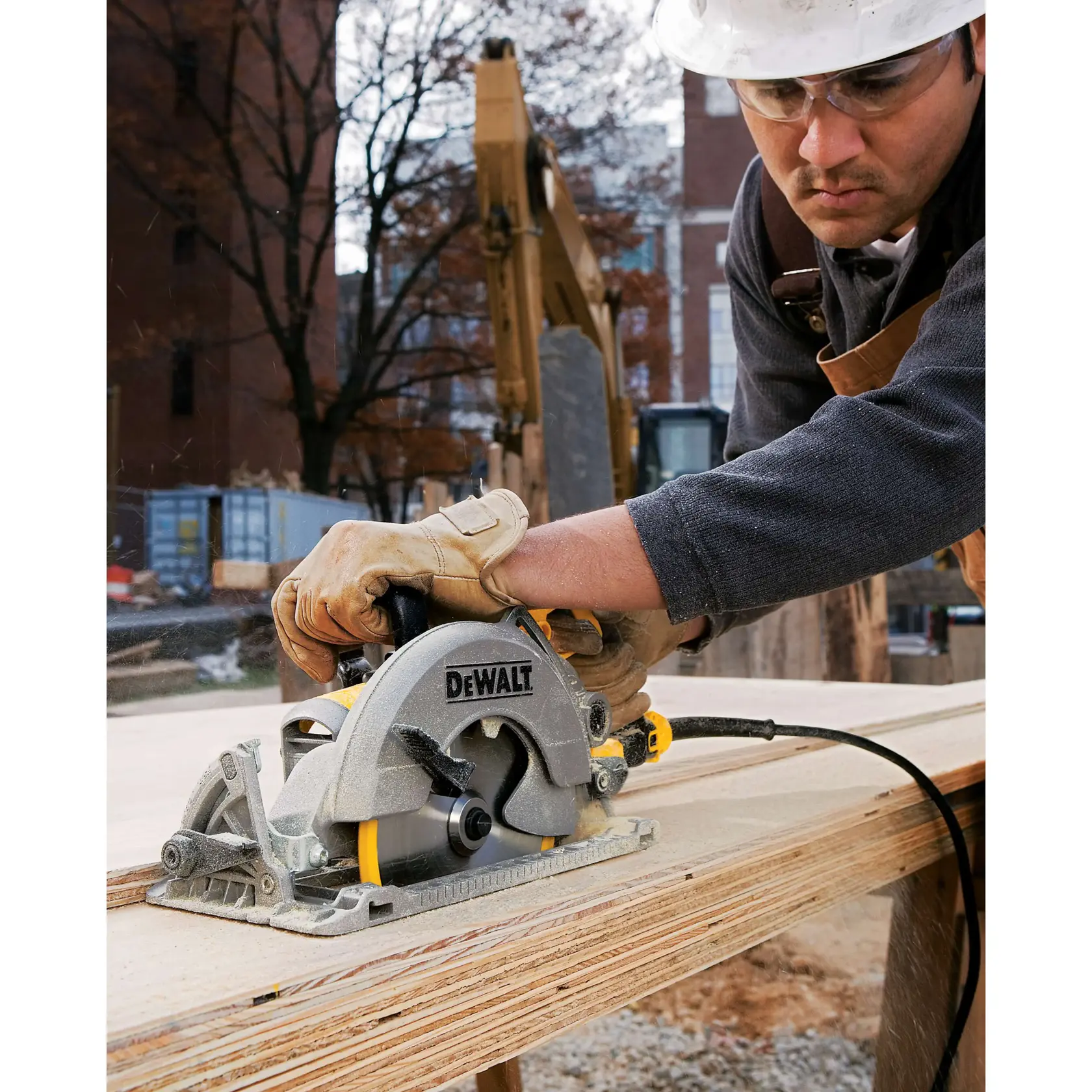 A person wearing protective gloves and a helmet is using a DEWALT circular saw to cut wood at an outdoor construction site. The saw is plugged in and there is construction equipment and buildings in the background.