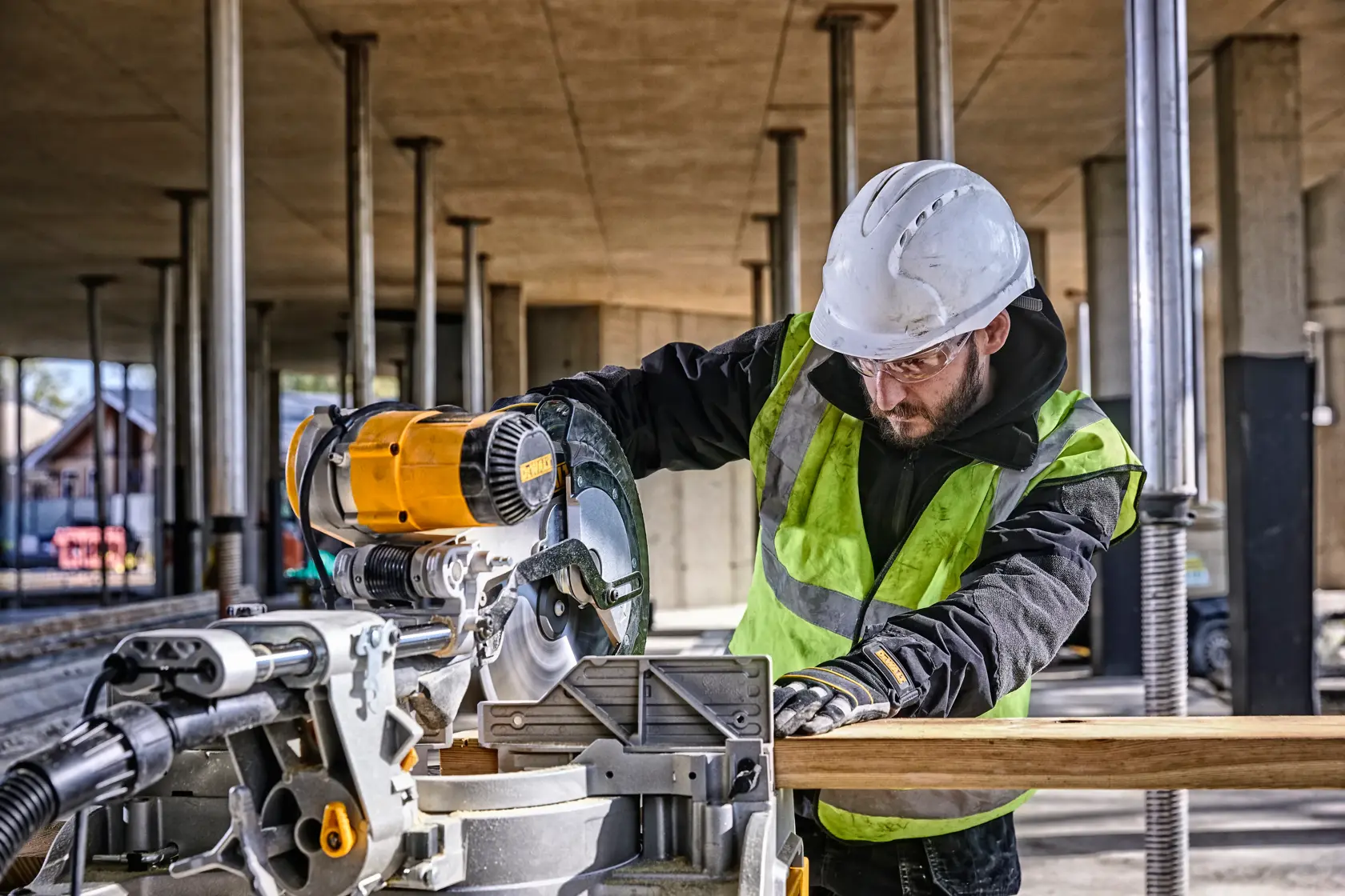 A construction worker wearing a safety vest and hard hat is using a DEWALT DWS780 miter saw to cut a piece of wood at an outdoor construction site.