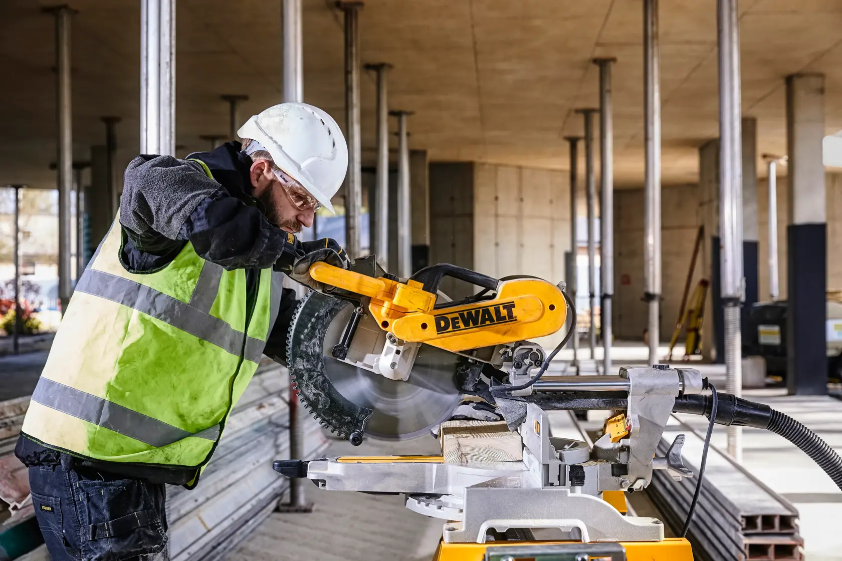 A construction worker wearing a safety vest and helmet operates a DEWALT DWS780 miter saw on a construction site. The worker's face is blurred for privacy.