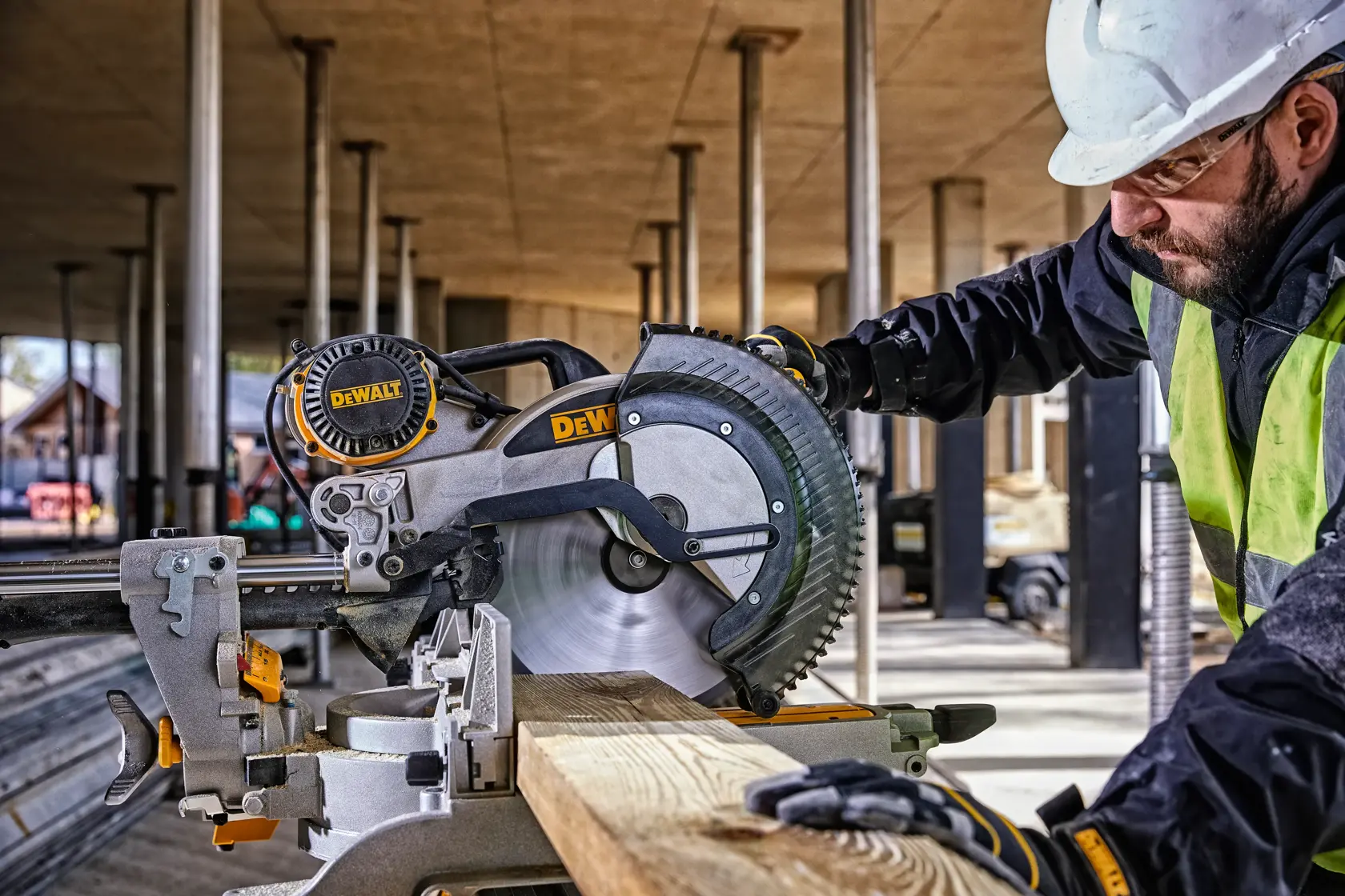 A DEWALT DWS780 miter saw is being operated by a worker wearing safety gear and a helmet at a construction site. The saw is cutting a piece of wood.