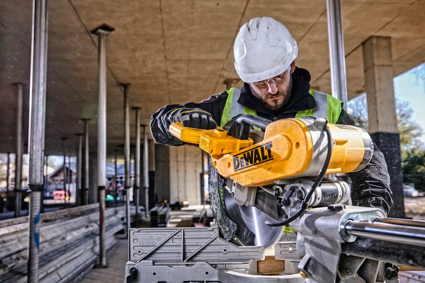 A person wearing a hard hat and safety vest is operating a DEWALT DWS780 miter saw at a construction site with metal supports and building materials visible in the background.