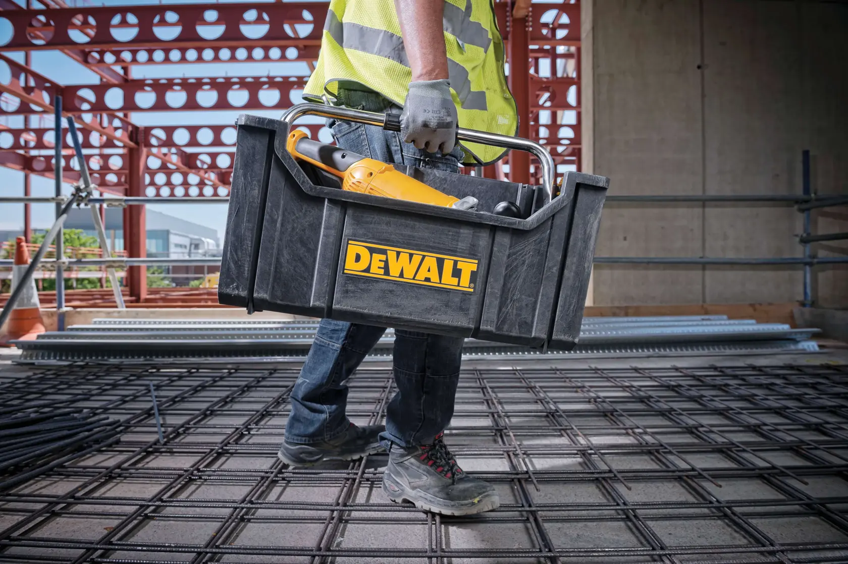 A construction worker carrying a black DEWALT toolbox with tools inside at a building site. Only the lower half of the person is visible, wearing work gloves, jeans, safety boots, and a high-visibility vest.