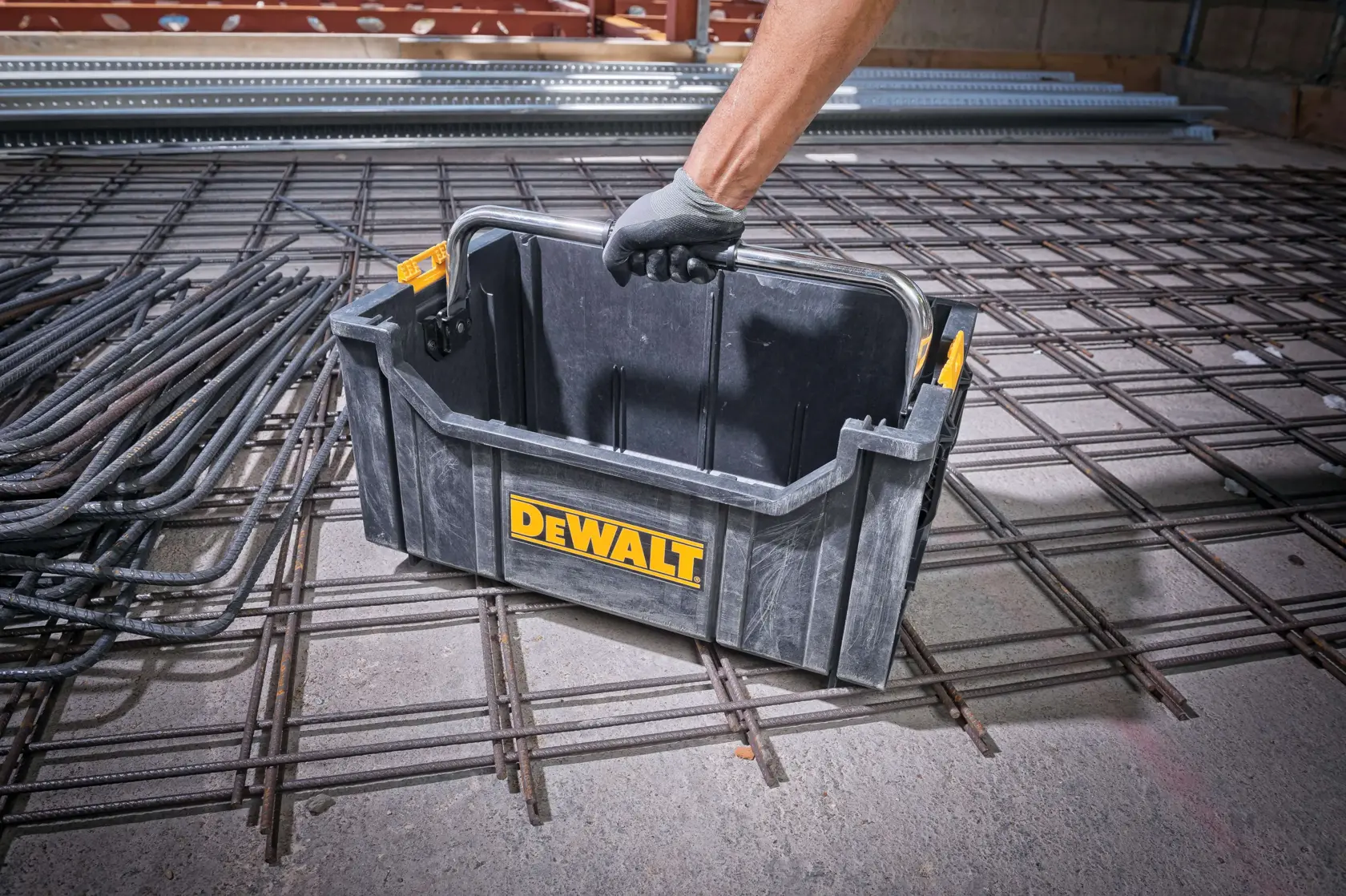 A DEWALT-branded black plastic toolbox with a metal handle is being carried by a person wearing a gray glove, positioned on a construction site with steel rebar and concrete in the background.