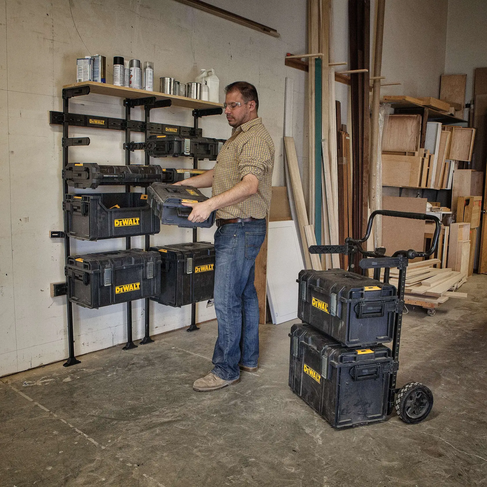 A person in a workshop organizes DEWALT storage boxes on a wall-mounted shelving system. More DEWALT storage boxes are stacked on a wheeled cart, surrounded by wood materials and shelves.