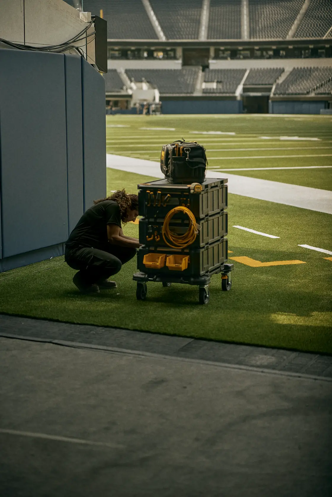 A person crouching next to a mobile DEWALT storage cart with stacked tool boxes and a coiled yellow cable, located on an indoor stadium field.