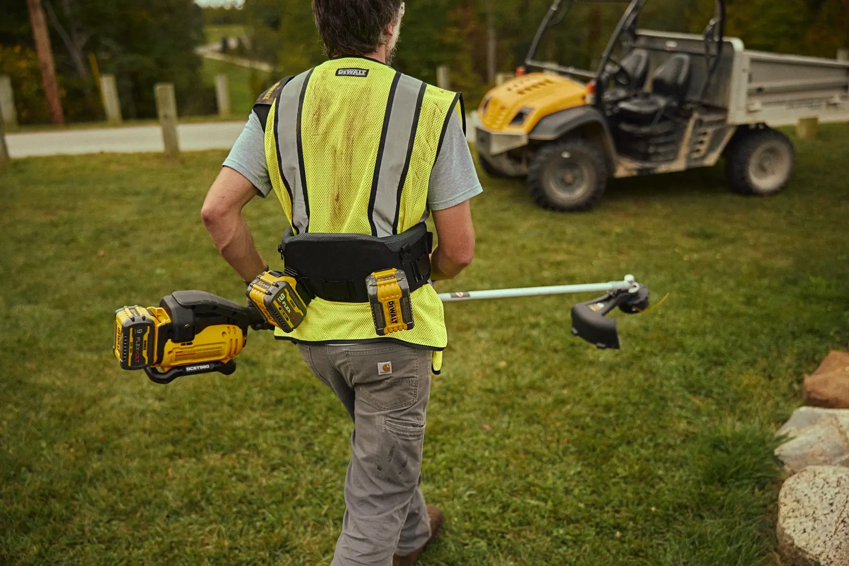 A person wearing a yellow safety vest is outdoors, carrying a DEWALT cordless string trimmer and wearing a DEWALT tool belt with batteries. In the background, there is a yellow utility vehicle on grass.