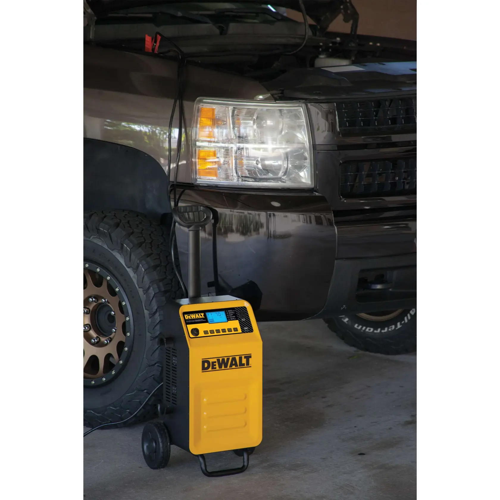 A DEWALT portable car battery charger (model DXAEC210) with wheels is connected to a black pickup truck in a garage. The truck's hood is open, and the charger is plugged into the vehicle battery.