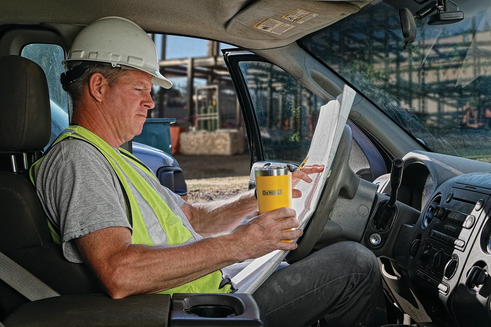 A person wearing a safety vest and hard hat sits in the driver's seat of a vehicle, holding a DEWALT-branded yellow insulated tumbler and reviewing paperwork or blueprints.