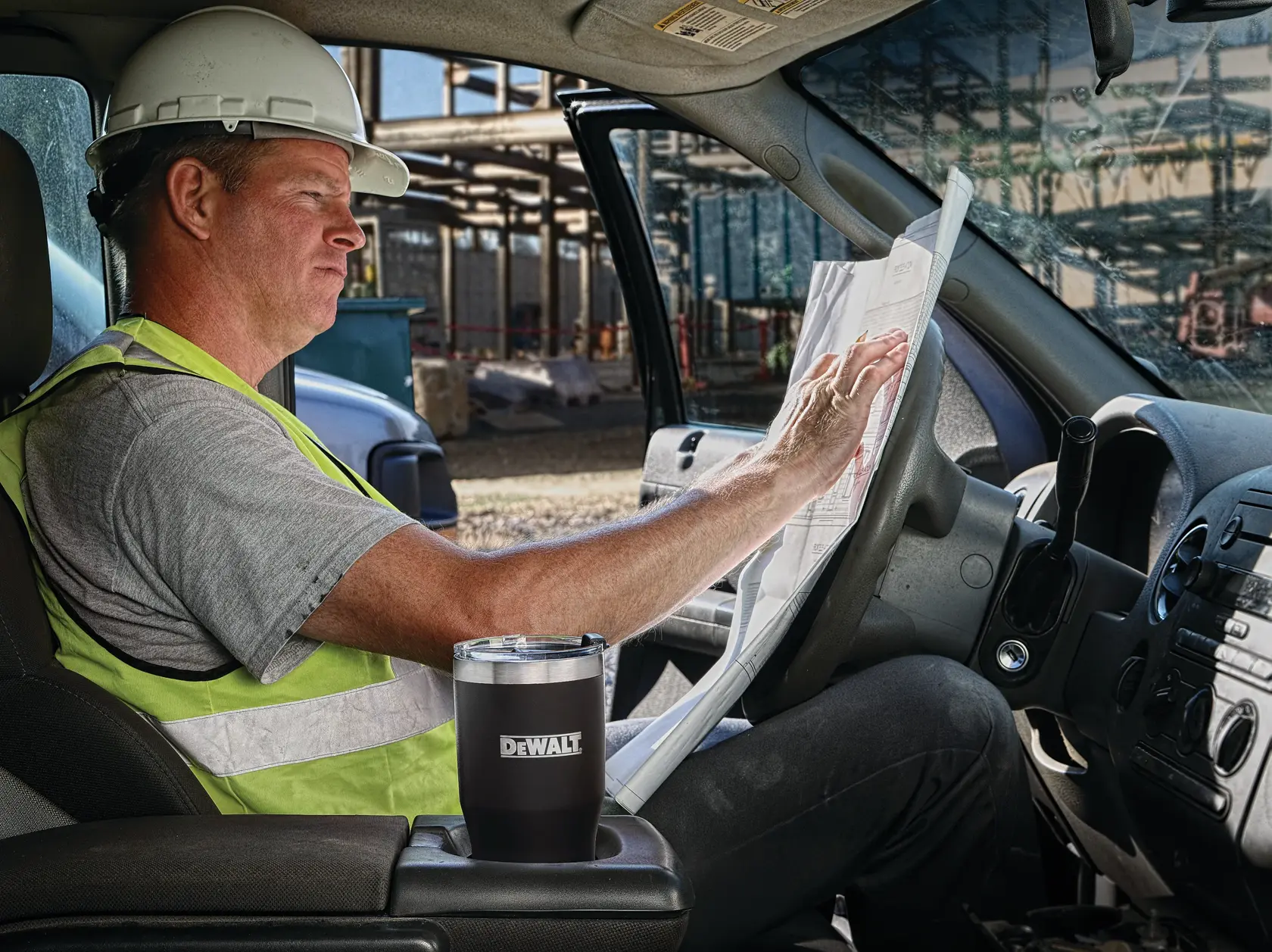 A worker wearing a safety vest and hard hat sits inside a vehicle on a construction site, holding a blueprint. A DEWALT branded tumbler is placed in the cup holder.
