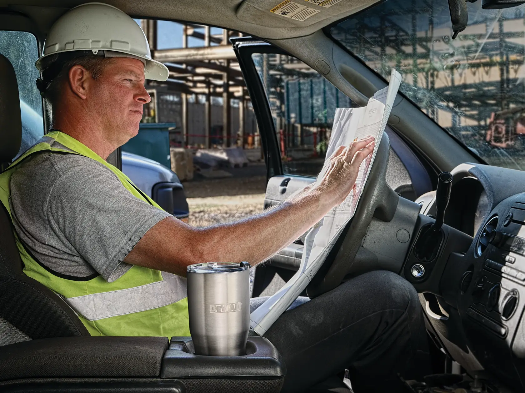 A person wearing a hard hat and safety vest sits inside a vehicle at a construction site, looking at blueprints. A stainless steel DEWALT travel tumbler is placed in the vehicle's cup holder.