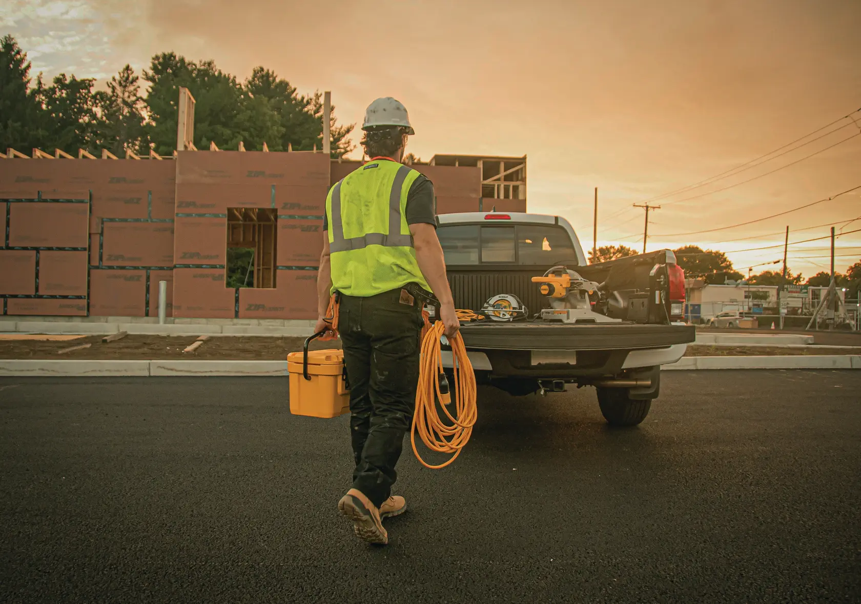 A construction worker wearing a safety vest and helmet walks towards a pickup truck parked in front of a building under construction, carrying a yellow tool box and a long orange extension cord.