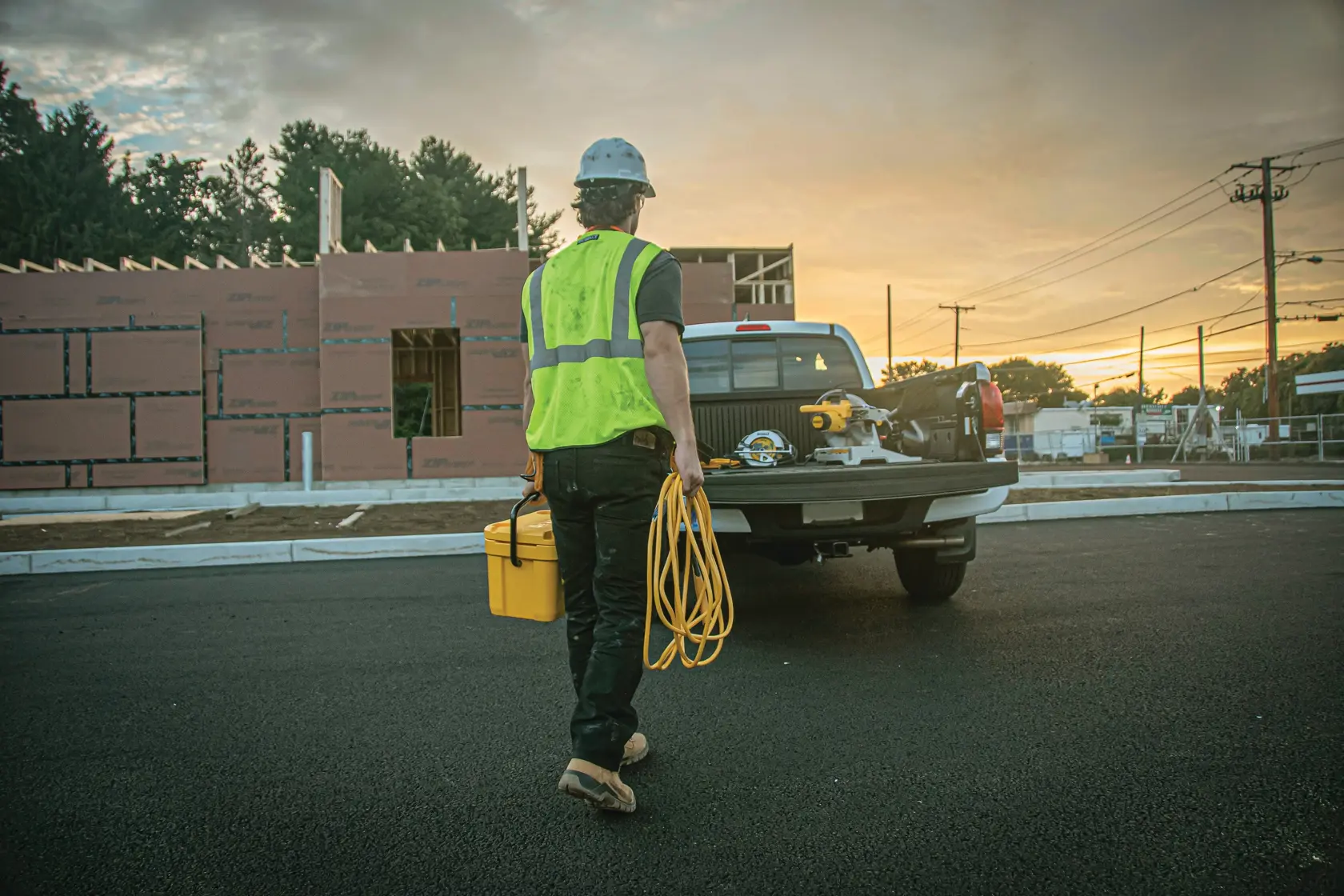 A construction worker wearing a reflective safety vest and helmet carries a yellow toolbox and a yellow extension cord towards the back of a pickup truck at a construction site during sunset.