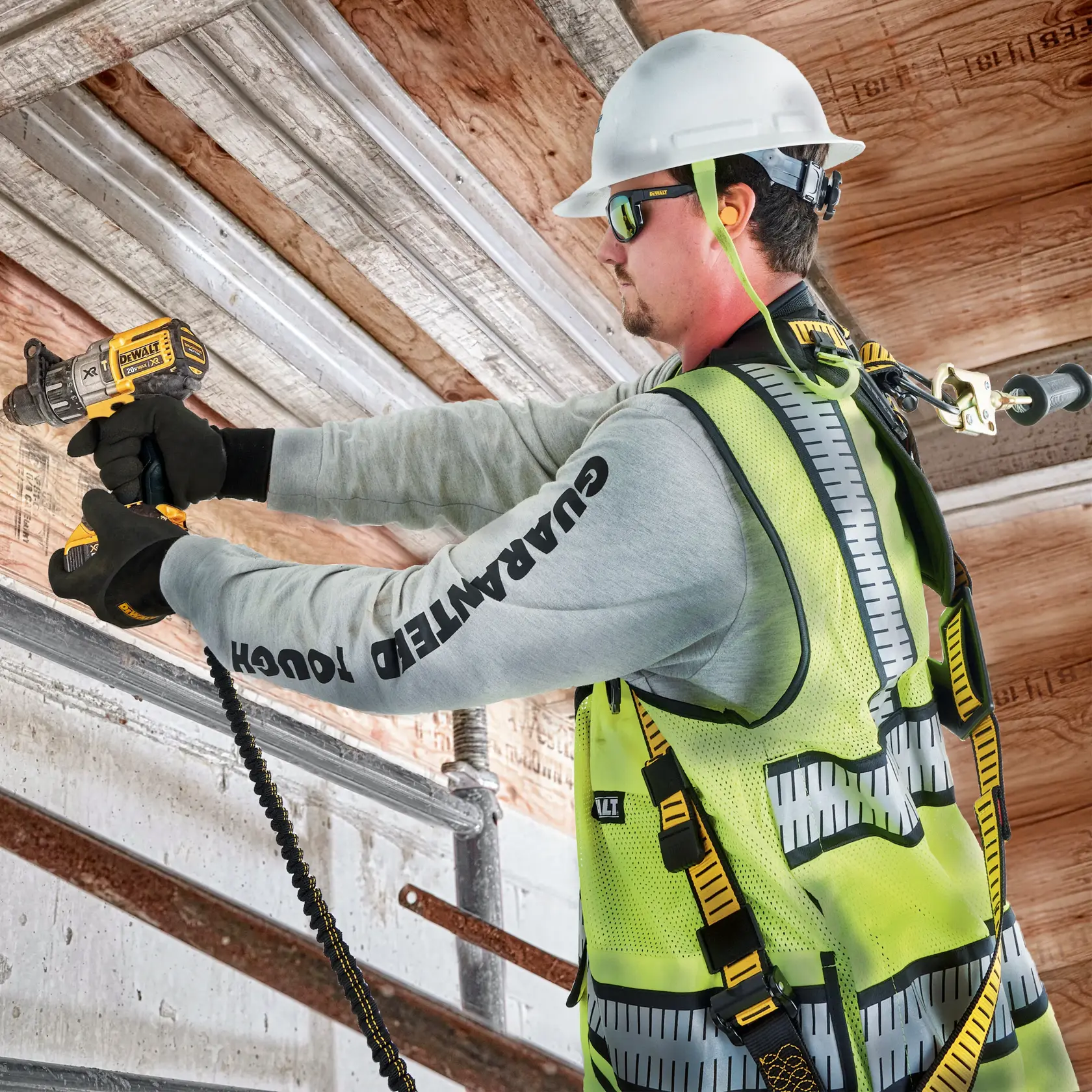 A construction worker wearing a safety helmet, high-visibility vest, and fall protection harness uses a DEWALT cordless drill to work on a metal beam. The worker's face is blurred for privacy.