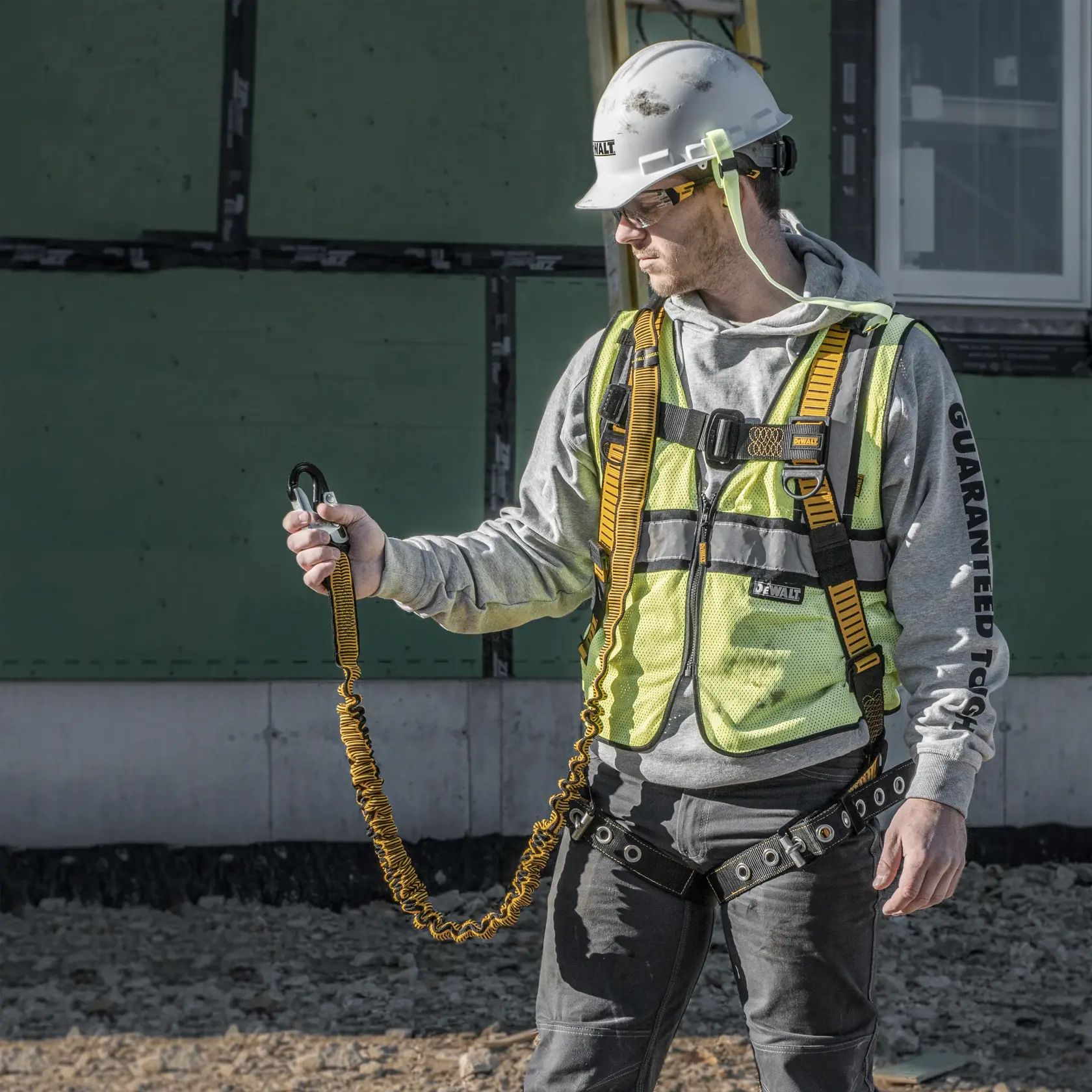A person wearing a DEWALT branded safety harness, high-visibility vest, and helmet is holding a yellow lanyard with a carabiner at a construction site. The person's face is blurred for privacy.